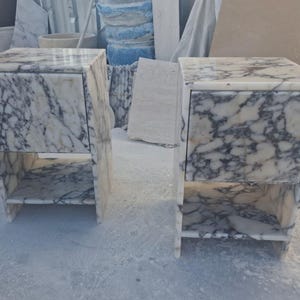 May include: Two marble nightstands with a white and gray veined pattern. Each nightstand has a drawer and an open shelf. The nightstands are rectangular and have a modern design. The nightstands are made of stone.
