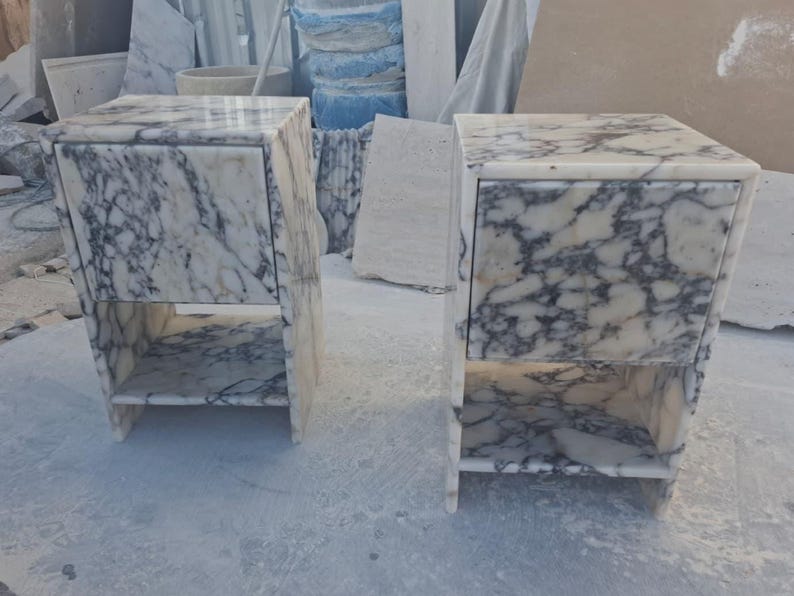 May include: Two marble nightstands with a white and gray veined pattern. Each nightstand has a drawer and an open shelf. The nightstands are rectangular and have a modern design. The nightstands are made of stone.