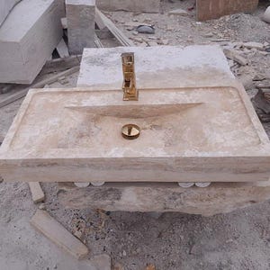May include: A rectangular beige travertine stone sink featuring a polished gold-tone faucet and a matching gold-tone drain cover. The basin has a smooth, flat rim and a sloped interior surface, showcasing the natural porous texture of the stone.