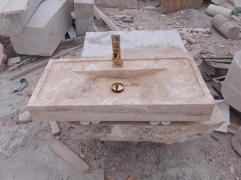 May include: A rectangular beige travertine stone sink featuring a polished gold-tone faucet and a matching gold-tone drain cover. The basin has a smooth, flat rim and a sloped interior surface, showcasing the natural porous texture of the stone.