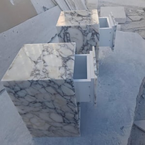 May include: Two marble nightstands with drawers. The nightstands are white with gray veining. One drawer is open, revealing a white interior. The nightstands are cube-shaped and sit on a light gray surface.