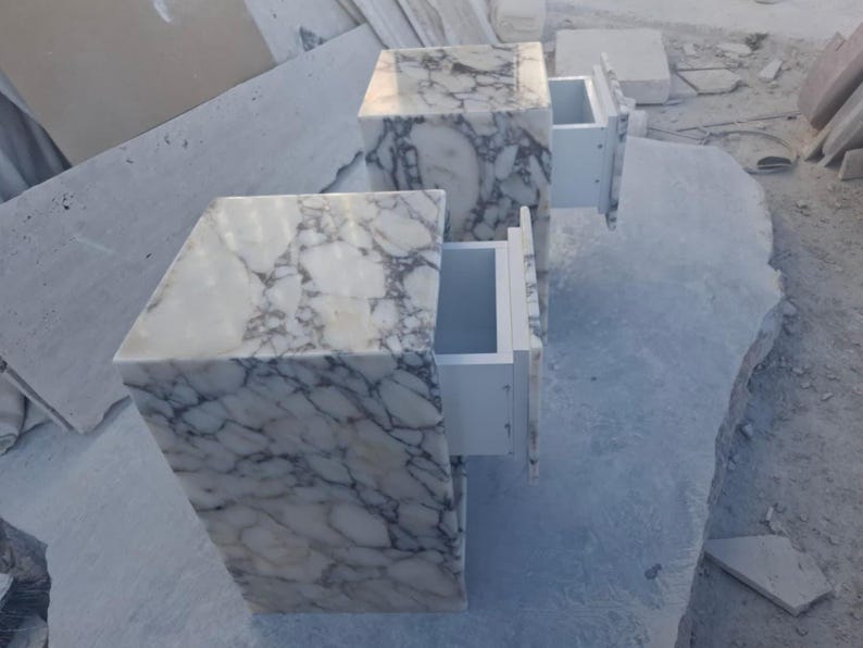 May include: Two marble nightstands with drawers. The nightstands are white with gray veining. One drawer is open, revealing a white interior. The nightstands are cube-shaped and sit on a light gray surface.