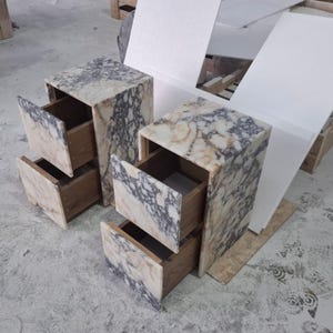 May include: Two bedside tables featuring a natural stone exterior with a marbled pattern in shades of cream, beige, and dark grey. Each table has two open drawers with visible wooden interiors, showcasing a boxy, rectangular construction.
