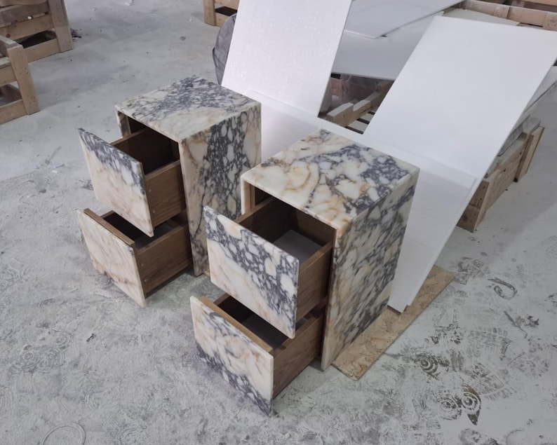May include: Two bedside tables featuring a natural stone exterior with a marbled pattern in shades of cream, beige, and dark grey. Each table has two open drawers with visible wooden interiors, showcasing a boxy, rectangular construction.