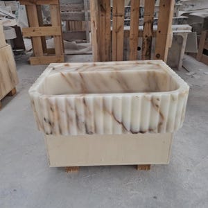 May include: A rectangular stone sink featuring a fluted exterior design with vertical grooves. The material is a cream-colored stone with prominent brown and grey veining throughout the basin and the outer edges, resting on a wooden base.