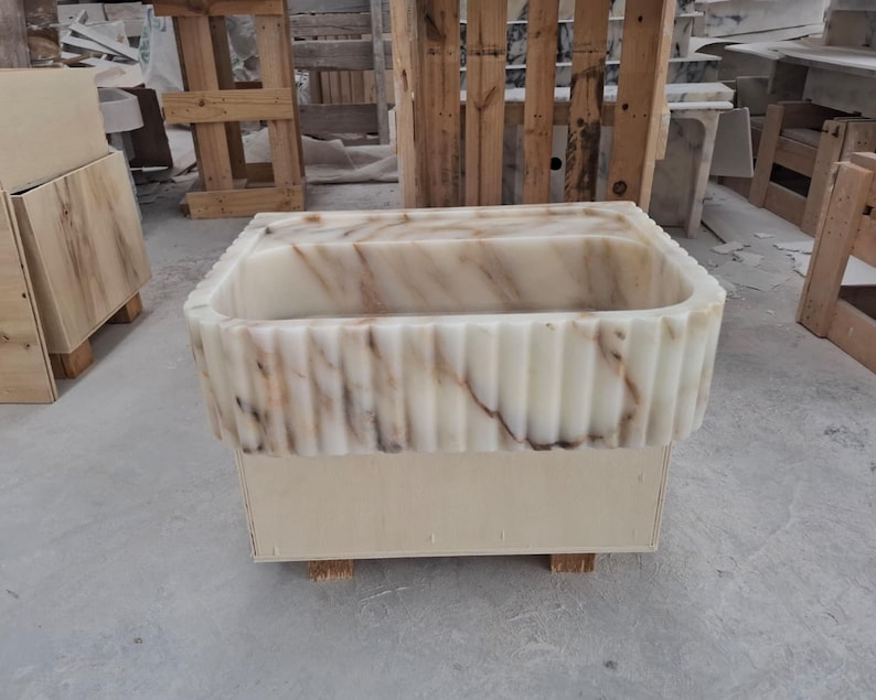 May include: A rectangular stone sink featuring a fluted exterior design with vertical grooves. The material is a cream-colored stone with prominent brown and grey veining throughout the basin and the outer edges, resting on a wooden base.