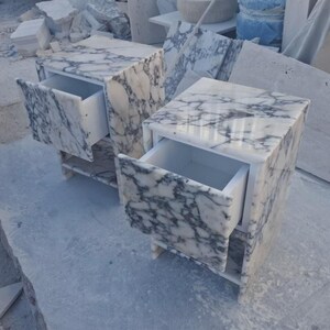 May include: Two marble nightstands with open drawers. The nightstands are white with gray veining, and have a rectangular shape. The drawers are white. The nightstands are sitting on a gray surface.