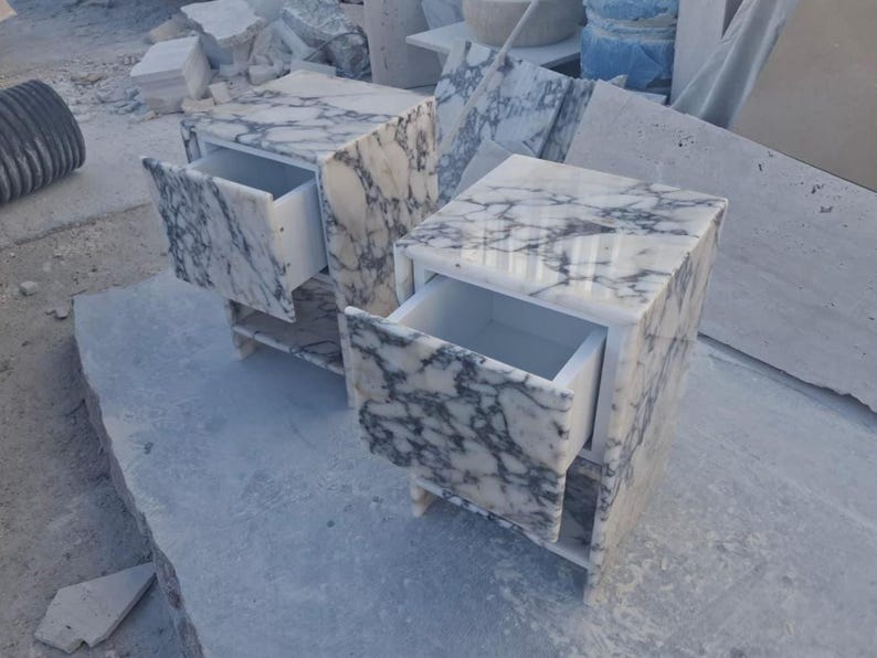 May include: Two marble nightstands with open drawers. The nightstands are white with gray veining, and have a rectangular shape. The drawers are white. The nightstands are sitting on a gray surface.