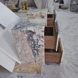 May include: A bedside table crafted from polished marble with a complex pattern of white, cream, and dark grey veining. The unit features three stacked drawers with wooden interiors and a matching marble front panel on each drawer face.