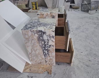 May include: Two marble-topped nightstands with wooden drawers. The marble has a mix of white, gray, and brown veining. The drawers are open, revealing the wooden interiors. The nightstands are in a workshop setting.