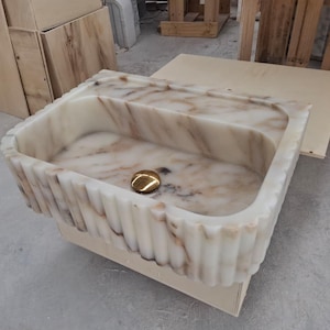 May include: A rectangular marble sink featuring a cream and brown veined pattern with a fluted exterior design. The basin includes a polished gold-toned circular drain cover centered at the bottom, showcasing a natural stone texture throughout the piece.