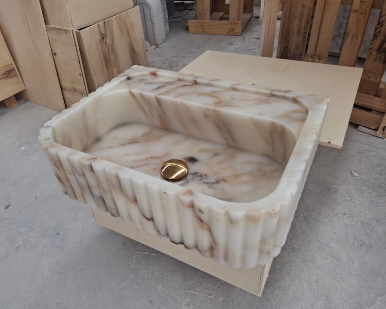 May include: A rectangular marble sink featuring a cream and brown veined pattern with a fluted exterior design. The basin includes a polished gold-toned circular drain cover centered at the bottom, showcasing a natural stone texture throughout the piece.