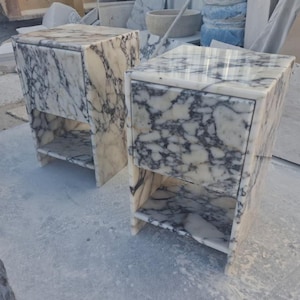 May include: Two marble nightstands with a white and gray veined pattern. Each nightstand has a drawer and an open shelf. The nightstands are rectangular and sit on small legs. The marble has a polished finish.