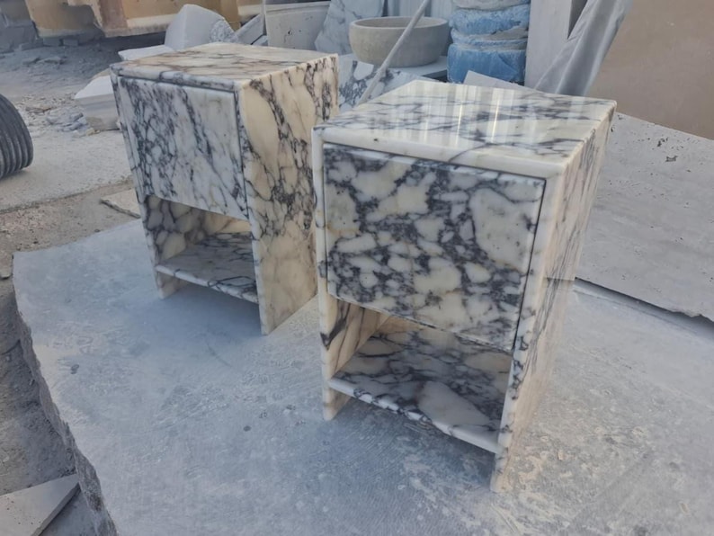 May include: Two marble nightstands with a white and gray veined pattern. Each nightstand has a drawer and an open shelf. The nightstands are rectangular and sit on small legs. The marble has a polished finish.