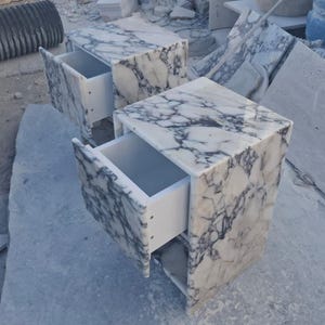 May include: Two marble nightstands with drawers open, showcasing a white interior. The marble has a white base with gray veining. The nightstands are cube-shaped and sit on a light gray surface.