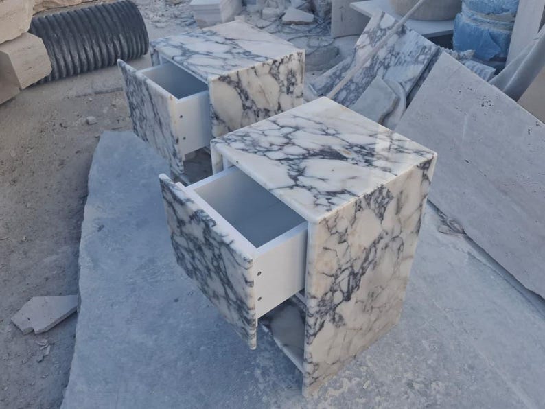 May include: Two marble nightstands with drawers open, showcasing a white interior. The marble has a white base with gray veining. The nightstands are cube-shaped and sit on a light gray surface.