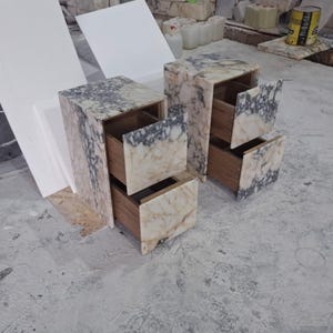 May include: Two bedside tables constructed from cream and dark grey veined marble. Each unit features a rectangular frame with two pull-out drawers. The drawer fronts are finished in matching marble, while the interior drawer boxes are made of wood.