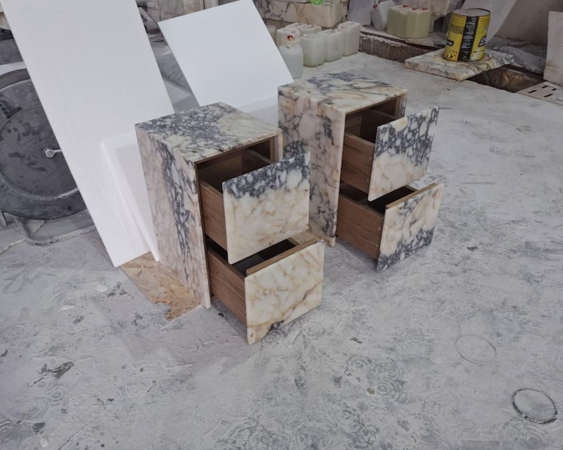 May include: Two bedside tables constructed from cream and dark grey veined marble. Each unit features a rectangular frame with two pull-out drawers. The drawer fronts are finished in matching marble, while the interior drawer boxes are made of wood.