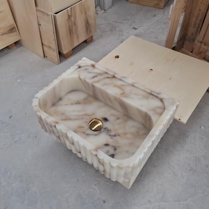 May include: A rectangular stone sink featuring a white and beige marbled pattern with a fluted exterior design. The basin includes a centered, polished gold-toned circular drain cover, showcasing a smooth interior finish and a textured outer edge.