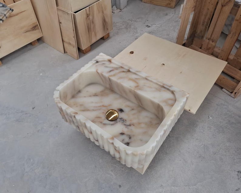 May include: A rectangular stone sink featuring a white and beige marbled pattern with a fluted exterior design. The basin includes a centered, polished gold-toned circular drain cover, showcasing a smooth interior finish and a textured outer edge.