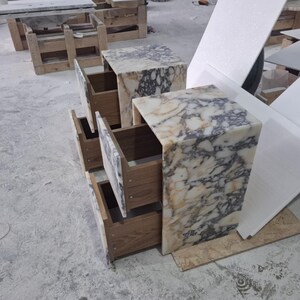 May include: Two bedside tables constructed from white and grey veined marble with open wooden drawers. The stone features a high-gloss finish with intricate natural patterns, while the interior of the drawers reveals a light brown wood grain texture.