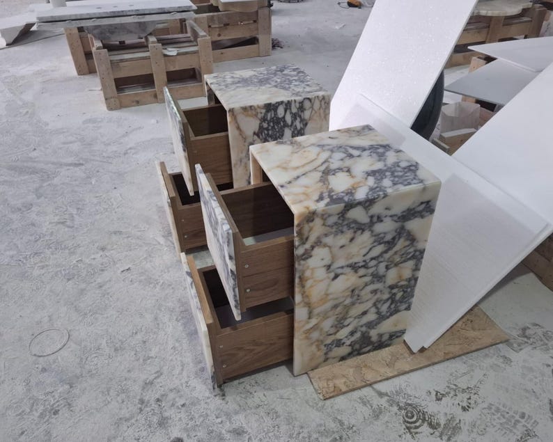 May include: Two bedside tables constructed from white and grey veined marble with open wooden drawers. The stone features a high-gloss finish with intricate natural patterns, while the interior of the drawers reveals a light brown wood grain texture.