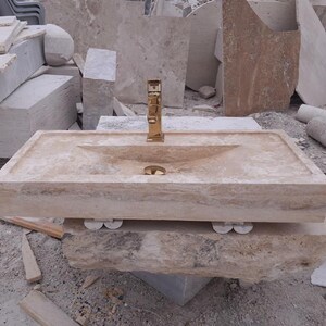 May include: A rectangular beige travertine stone sink featuring a polished gold-tone faucet centered at the back. The basin has a smooth, sloped interior design with a matching gold-tone drain cover, supported by four small, rounded stone feet underneath.