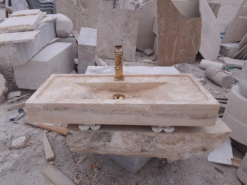 May include: A rectangular beige travertine stone sink featuring a polished gold-tone faucet centered at the back. The basin has a smooth, sloped interior design with a matching gold-tone drain cover, supported by four small, rounded stone feet underneath.