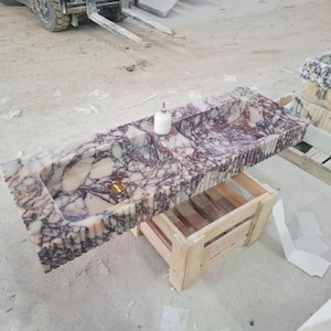 Peut inclure: Un évier rectangulaire en marbre avec un motif de couleurs blanches, violettes et bordeaux. L'évier a deux vasques, un drain doré et un distributeur de savon. Il repose sur une caisse en bois.