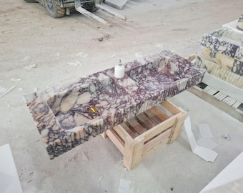 May include: A rectangular marble sink with a patterned design in shades of white, purple, and burgundy. The sink has two basins, a gold-colored drain, and a soap dispenser. It is resting on a wooden crate.