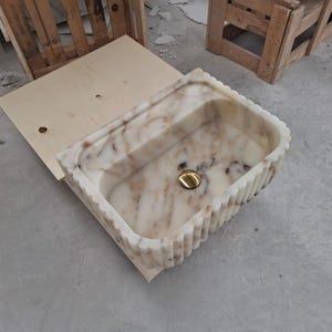 May include: A rectangular marble sink featuring a cream and brown veined pattern with a fluted exterior design. The basin includes a polished gold-toned circular drain cover centered at the bottom, showcasing a natural stone texture and finish.