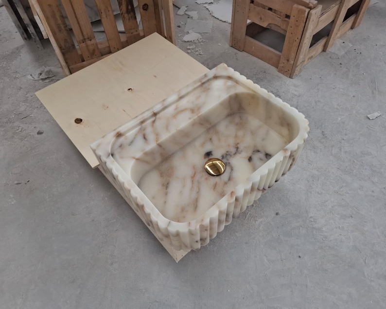 May include: A rectangular marble sink featuring a cream and brown veined pattern with a fluted exterior design. The basin includes a polished gold-toned circular drain cover centered at the bottom, showcasing a natural stone texture and finish.
