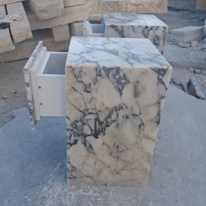 May include: Two marble bedside tables with a white and gray veined pattern. One drawer is open, revealing a white interior. The tables are rectangular with a square top and are set in a stone yard.