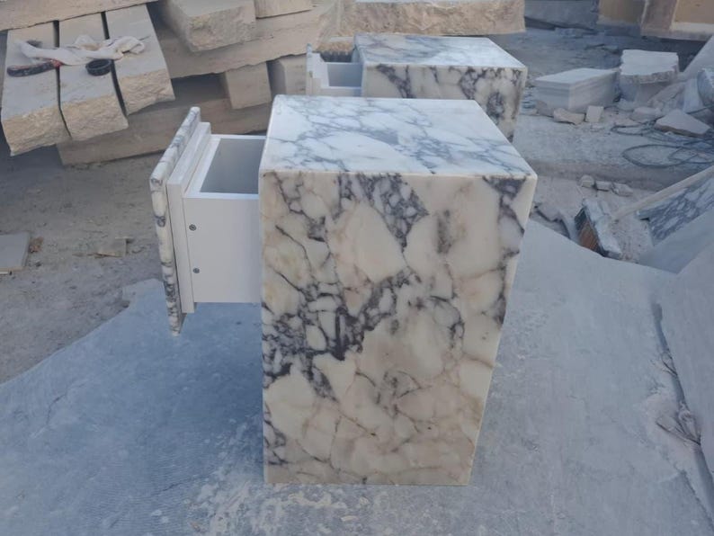 May include: Two marble bedside tables with a white and gray veined pattern. One drawer is open, revealing a white interior. The tables are rectangular with a square top and are set in a stone yard.