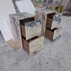 May include: Two bedside tables constructed from white and grey veined marble with two drawers each. The drawer fronts are faced with matching marble slabs, while the interior drawer boxes are made of brown wood, visible as they are pulled open.