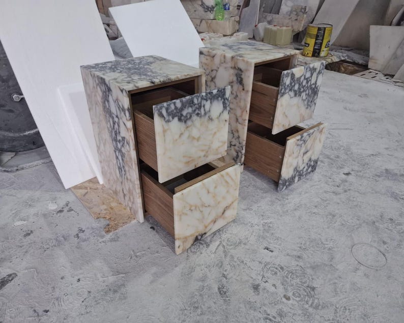 May include: Two bedside tables constructed from white and grey veined marble with two drawers each. The drawer fronts are faced with matching marble slabs, while the interior drawer boxes are made of brown wood, visible as they are pulled open.