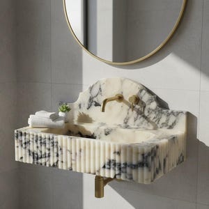 May include: A marble washbasin with a gold tap and waste pipe. The basin features a white and grey veined pattern and a fluted front. A gold-framed mirror hangs above, and a small plant and folded towels sit on the edge of the basin.