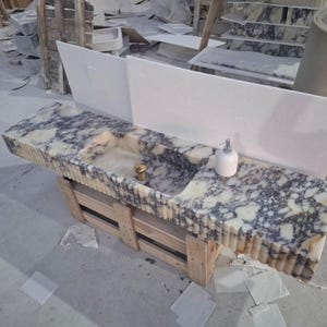 May include: A long rectangular vanity countertop and integrated sink made of white and dark gray marbled stone. The front edge features a vertical fluted design. A white soap dispenser and a gold-toned drain fixture are placed on the surface.