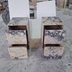 May include: Two matching bedside tables crafted from white and grey veined marble. Each unit features a rectangular frame with two stacked drawers, showcasing a continuous natural stone pattern across the front panels and the top surface.