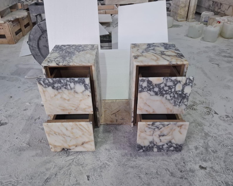 May include: Two matching bedside tables crafted from white and grey veined marble. Each unit features a rectangular frame with two stacked drawers, showcasing a continuous natural stone pattern across the front panels and the top surface.