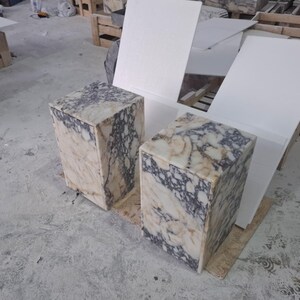 May include: Two rectangular side tables crafted from polished marble featuring a distinct veined pattern in shades of cream, beige, and deep purple. Each table has a solid block construction with a smooth top surface and vertical sides.