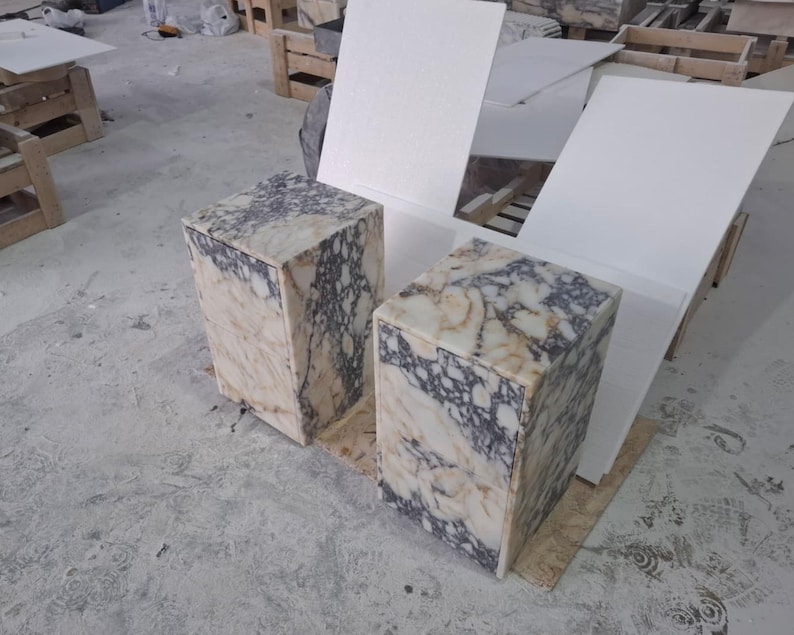 May include: Two rectangular side tables crafted from polished marble featuring a distinct veined pattern in shades of cream, beige, and deep purple. Each table has a solid block construction with a smooth top surface and vertical sides.