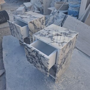 May include: Two marble nightstands with drawers. The nightstands are white with gray veining. The drawers are open, revealing the white interior. The nightstands are square and sit on a light gray surface.