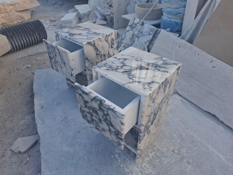 May include: Two marble nightstands with drawers. The nightstands are white with gray veining. The drawers are open, revealing the white interior. The nightstands are square and sit on a light gray surface.