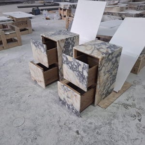 May include: Two bedside tables featuring a natural stone exterior with a marbled pattern of cream, beige, and dark grey veins. Each unit has two drawers with wooden interiors and a rectangular frame constructed from the same stone material.