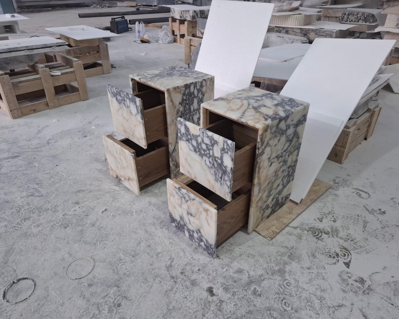May include: Two bedside tables featuring a natural stone exterior with a marbled pattern of cream, beige, and dark grey veins. Each unit has two drawers with wooden interiors and a rectangular frame constructed from the same stone material.