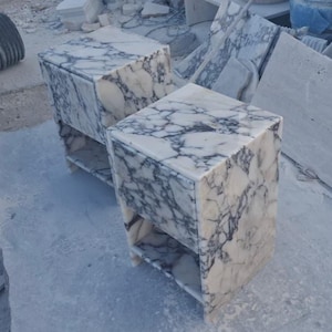 May include: Two marble nightstands with a white and gray veined pattern. Each nightstand has a single shelf and a cube shape. The nightstands are made of stone and are sitting on a concrete surface.
