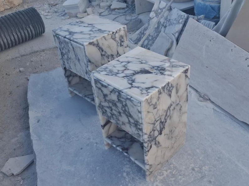 May include: Two marble nightstands with a white and gray veined pattern. Each nightstand has a single shelf and a cube shape. The nightstands are made of stone and are sitting on a concrete surface.