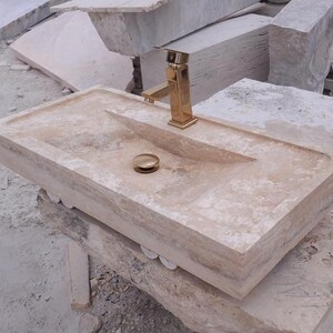 May include: A rectangular beige travertine stone sink featuring a polished gold-tone faucet and a matching gold-tone drain stopper. The basin has a smooth, flat rim and a sloped interior surface, showcasing the natural porous texture of the stone.
