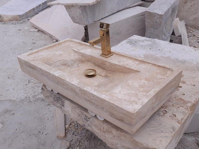May include: A rectangular beige travertine stone sink featuring a polished gold-tone faucet and a matching gold-tone drain stopper. The basin has a smooth, flat rim and a sloped interior surface, showcasing the natural porous texture of the stone.